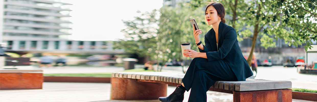 Nữ doanh nhân trẻ sử dụng điện thoại thông minh trên băng ghế ở công viên với tách cà phê trên tay, hình ảnh sử dụng cho trang tính năng Digital Secure Key của HSBC Việt Nam